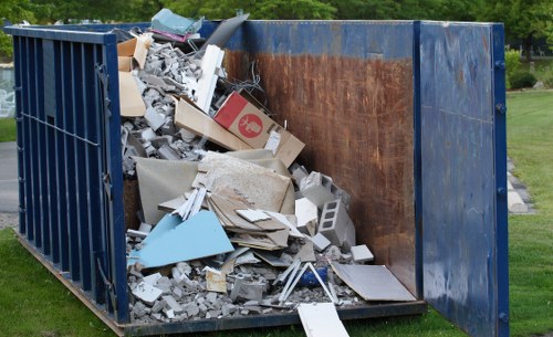 An open-top, large industrial waste skip situated outdoors on a grassy area, filled with a mix of construction and household debris including various broken concrete pieces, shattered tiles, fragments of drywall, cardboard boxes, and discarded wooden and plastic materials. The skip's sides are made of dark blue metal with a rusted, weathered interior surface visible from the open top. Surrounding the skip, there is a green lawn and some blurred trees in the background, indicating a suburban or park-like setting. The waste appears haphazardly piled, with some items spilling over the edges, consistent with rubbish removal services provided by Chelsea House Clearance in the Chelsea or surrounding areas, including postal codes within London. The scene is lit by natural daylight, emphasizing the textures and colors of the diverse waste materials.