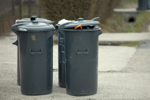 Two dark grey plastic wheelie bins with hinged lids are positioned on a paved surface, likely a driveway or street edge. The bin on the left has its lid closed, while the one on the right has an open lid revealing a partially visible orange object or tool inside. Both bins are standard size, with a slightly textured surface and rectangular handles on the front. The background shows a blurred outdoor scene with trees, greenery, and a distant footpath or road, suggesting a residential area in Chelsea or nearby. The lighting indicates daytime with natural light illuminating the scene evenly. The image relates to rubbish collection or waste disposal services provided by Chelsea House Clearance, with the bins designated for general waste. The setting appears orderly with no visible debris or litter around the bins, emphasizing proper waste management practices in an urban neighborhood.