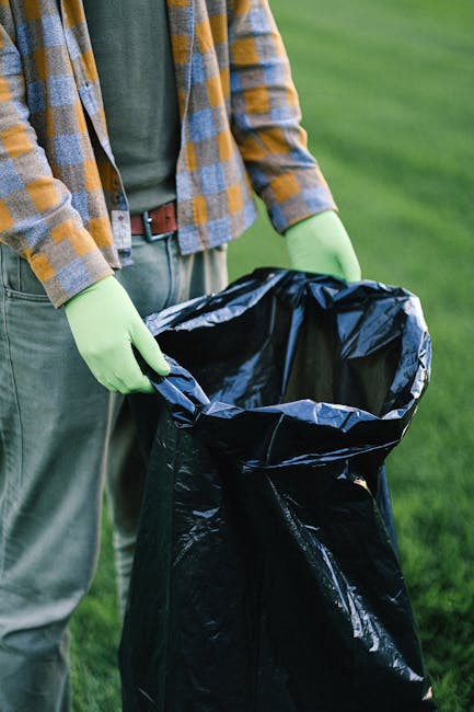 An overhead view of a female street cleaner dressed in bright orange high-visibility overalls and a matching orange cap, standing on a grey pavement. She is holding a broom with a wooden handle and re